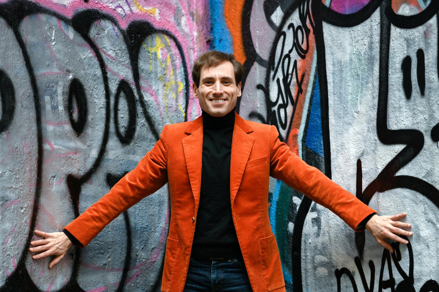 Boris Giltburg in orange jacket, standing in front of a wall covered with colourful graffiti